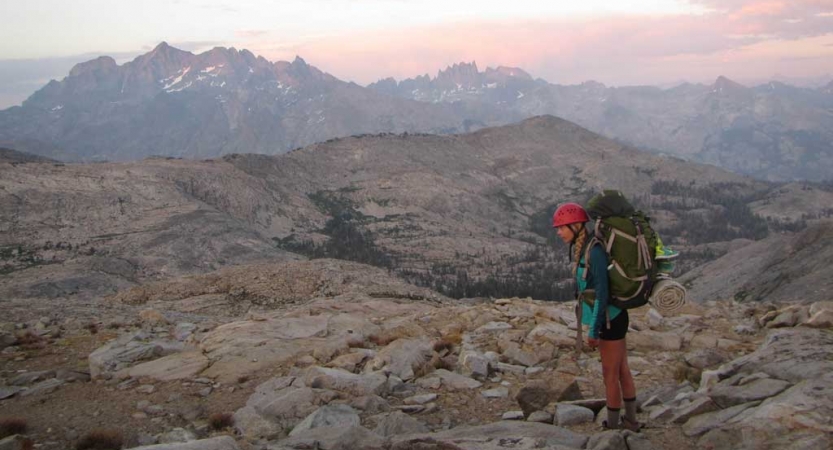 A person wearing a backpack stands in a rocky landscape with mountains in the background. The sky appears in shades of pink. 